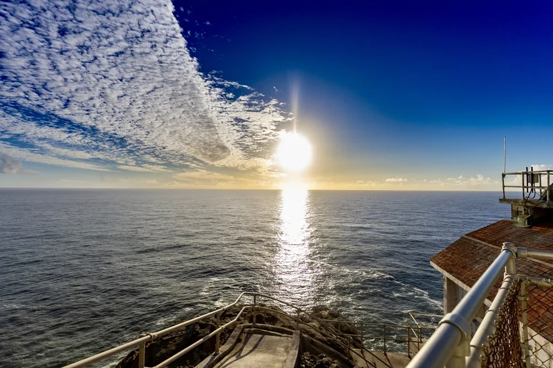 Sunlight reflects across the water beside a waterfront railing, beneath streaked clouds.